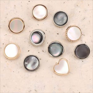 Può includere: Una collezione di bottoni decorativi con centri in madreperla iridescente. I bottoni hanno bordi in metallo dorato, argentato e nero, incluso un bottone a forma di cuore. Sono disposti su una superficie beige chiaro.