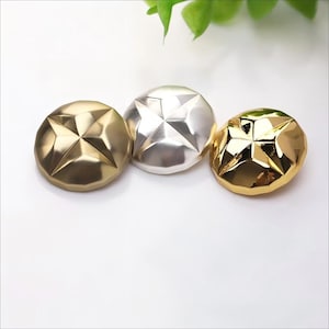 May include: Three metallic buttons in gold, silver, and bronze. Each button has a starburst design in the center. The buttons are round and have a faceted surface, reflecting light. The buttons are likely for sewing or crafts.