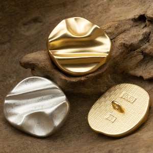 以下が含まれることがあります： 異なるデザインの金色のボタンが3つあります。1つのボタンは波打った質感のある表面をしています。もう1つのボタンはグリッドパターンです。3つ目のボタンは滑らかで波打った表面をしています。