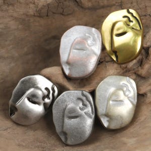 Puede incluir: Cinco botones de metal en diferentes colores, cada uno con una cara estilizada con los ojos cerrados y el pelo ondulado. Los botones están dispuestos en círculo sobre una superficie de madera.