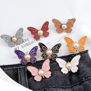May include: A collection of butterfly-shaped brooches in various colors, including blue, red, brown, orange, black, purple, pink, and white. Each brooch features a pearl in the center and intricate wing detailing. The text "YOU ARE MY SUNSHINE" is visible.