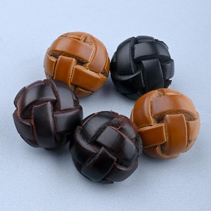 May include: Five leather buttons in shades of brown and black. Each button features a woven design, with leather strips interlaced to form a textured sphere. The buttons are displayed on a light blue surface.