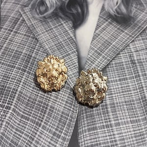 Puede incluir: Dos broches de cabeza de león de color dorado, uno con un acabado más pulido y el otro con un acabado más texturizado, están prendidos a una chaqueta gris a cuadros.