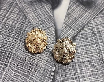 May include: Two gold-toned lion head brooches, one with a more polished finish and the other with a more textured finish, are pinned to a grey plaid blazer.