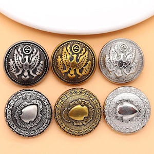 Puede incluir: Seis botones de metal decorativos con diseños ornamentados. Dos botones presentan un diseño de águila dorada, dos botones presentan un diseño de escudo dorado y dos botones presentan un diseño de escudo plateado.