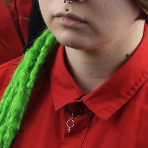 May include: A person wearing a red shirt with a green and black braided hairpiece. The person is wearing a silver necklace with a female symbol pendant.