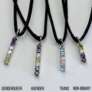 Puede incluir: Cuatro collares con colgantes de plata y cordón negro. Cada colgante es una barra vertical con gemas de colores. El texto en la parte inferior dice: GENDERQUEER, AGENDER, TRANS, NON-BINARY.