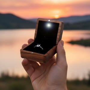 May include: A wooden ring box with a black velvet interior, lit by a small light. The box is open, revealing a silver engagement ring. The background shows a sunset over a lake and mountains.
