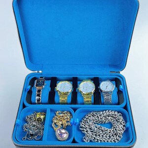 May include: Black leather watch box with blue velvet lining. The box has five slots for watches and three compartments for jewelry. The compartments contain a silver chain, a gold chain, and a pink crystal pendant.