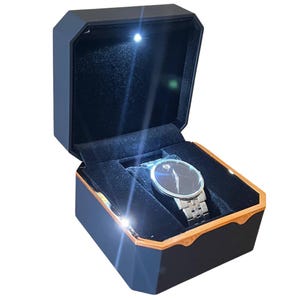 Könnte beinhalten: Eine schwarze Uhr mit silbernem Armband liegt in einer schwarzen Uhrenbox mit goldenem Rand. Die Box ist geöffnet und zeigt ein schwarzes Samtinterieur und ein kleines Licht, das die Uhr beleuchtet.