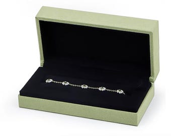 Könnte beinhalten: Eine grüne Samt-Schmuckschatulle mit schwarzem Samt-Innenfutter. Ein zartes silbernes Armband mit kleinen weißen und grünen Blumen-Anhängern ist in der Schachtel ausgestellt.