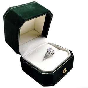 May include: A green velvet ring box with a white interior. A silver ring with a large, clear stone is displayed inside the box.