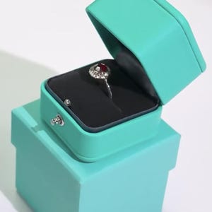 May include: A turquoise ring box with a red gemstone ring inside. The box is open and the ring is on a black velvet cushion.