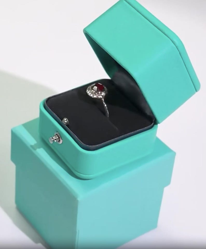 May include: A turquoise ring box with a red gemstone ring inside. The box is open and the ring is on a black velvet cushion.