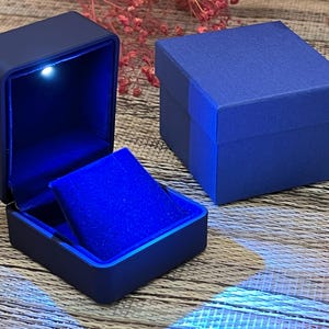 May include: Two blue jewelry boxes, one open and one closed. The open box has a bright blue velvet interior and a small light. The closed box is a solid blue. The boxes are on a wooden surface.