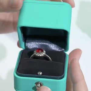 May include: A turquoise ring box with a silver ring with a red gemstone and clear stones. The box is open and the ring is inside.