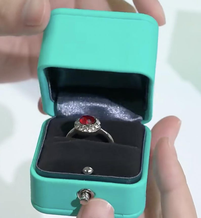 May include: A turquoise ring box with a silver ring with a red gemstone and clear stones. The box is open and the ring is inside.
