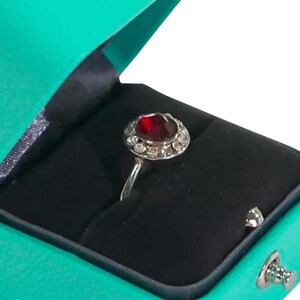 May include: A red gemstone ring with a silver band and a halo of smaller gemstones. The ring is in a teal velvet ring box.