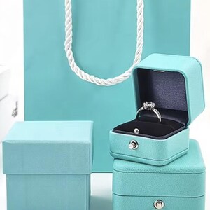 May include: A set of three turquoise blue ring boxes. The top box has a ring inside. The boxes are made of a textured material and have a silver button on the front.