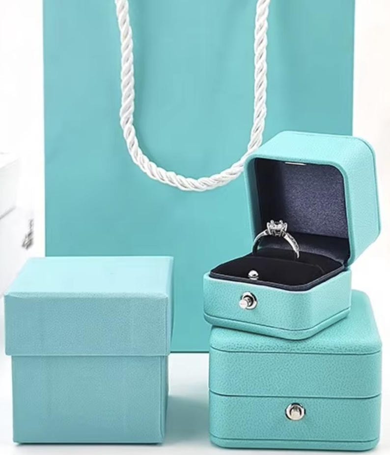 May include: A set of three turquoise blue ring boxes. The top box has a ring inside. The boxes are made of a textured material and have a silver button on the front.