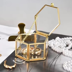 Puede incluir: Una joyero hexagonal de vidrio y oro con tapa con bisagras, que contiene dos anillos de oro. La caja está abierta, revelando los anillos en su interior. Una cadena dorada conecta la tapa a la base. Un clip dorado y un collar de perlas están en el fondo.