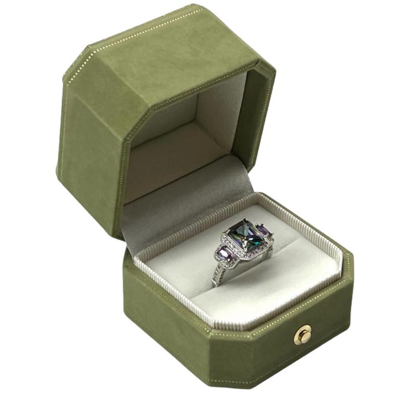 May include: A green velvet ring box with a white interior. A silver ring with a large, multi-colored gemstone is displayed inside the box.