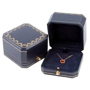 Pode incluir: Uma caixa de joias octogonal azul-escura com acabamento decorativo dourado e um pequeno fecho de cristal. A caixa aberta revela um colar de corrente dourada com um pequeno pingente em forma de coração vermelho sobre um forro de veludo azul.