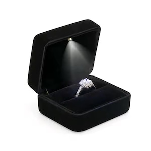 May include: A black velvet ring box with an integrated spotlight highlighting a diamond ring. The ring has a large, square-cut central stone and a band with smaller accent stones. The box is open, displaying the ring against a dark interior.