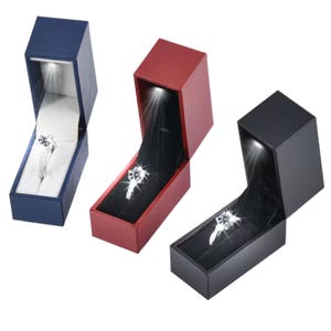 May include: Three rectangular jewellery boxes in blue, red, and black, each featuring an integrated LED light that illuminates a silver ring with a clear stone set inside. The boxes have a textured exterior finish and a hinged lid design.