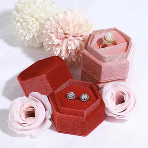 May include: A collection of velvet jewelry boxes in shades of red and pink, some open to display a ring and earrings. The ring features a pearl and gold setting. Artificial flowers in pink and white are arranged around the boxes.