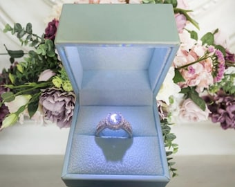 Könnte beinhalten: Eine hellblaue Ringbox mit weißem Innenraum und einem silbernen Ring mit einem großen, klaren Stein in der Mitte.