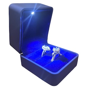 May include: A dark blue ring box with a built-in LED light illuminating two diamond rings. The interior is lined with blue velvet. The box is open, showcasing the rings in their individual compartments.