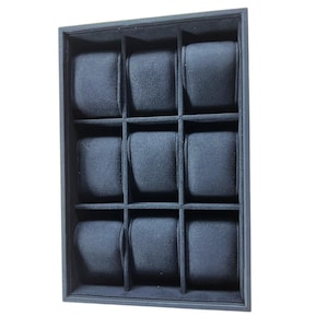 May include: A black watch display case with nine compartments. Each compartment has a soft, dark grey cushion to hold a watch. The case is rectangular and has a simple, clean design.