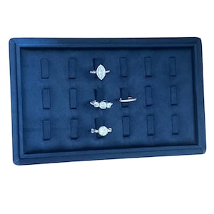 May include: A navy blue jewelry display tray with multiple slots, showcasing four diamond rings. The rings feature various designs, including a marquise cut, three-stone, and a round-cut diamond. The tray is rectangular with a raised border.