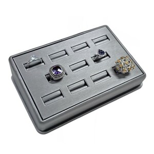 May include: A gray ring display tray with 12 slots for rings. The tray has a textured surface and is made of a sturdy material. There are four rings in the slots, including a silver ring with a clear stone, a silver ring with a purple stone, a silver ring with a blue stone, and a silver ring with a gold and white design.
