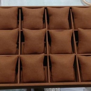 May include: A brown wooden watch display case with twelve individual compartments, each containing a brown, square pillow. The case is designed to hold and showcase watches, providing a neat and organized presentation.