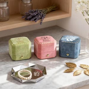 Könnte beinhalten: Drei Ringboxen aus Samt in Grün, Rosa und Blau, jede mit einem goldenen Knopf und personalisiertem Text oben. Ein silbernes geometrisches Tablett enthält ein Perlenarmband und zwei Ringe. Lavendel und dekorative Blätter im Hintergrund.