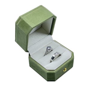 May include: Open green velvet ring box with two rings inside. One ring has a large, round, clear stone. The other ring is silver with a rectangular design. The box is octagonal with a gold clasp.