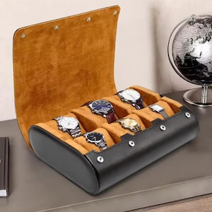 Pode incluir: Um estojo de relógio de couro preto com interior de camurça castanha, contendo vários relógios de pulso. O estojo está aberto, revelando os relógios com várias cores de pulseira e designs de mostrador. Um globo é visível no fundo.