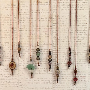 May include: A collection of eleven vertical hanging ornaments, each featuring a thin brown cord adorned with various beads, including clear, translucent, metallic, and carved stone elements, such as a green bear-shaped pendant.