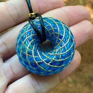 May include: A blue and gold geometric pendant necklace with a black cord. The pendant is a torus shape with a detailed, intricate design.