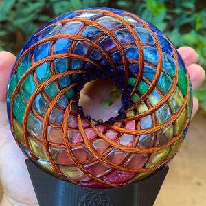 May include: A toroidal sculpture featuring a rainbow gradient of embedded colorful glass fragments arranged in a spiral pattern, wrapped in a dense copper wire coil, and mounted on a black stand with a circular geometric emblem on the front.