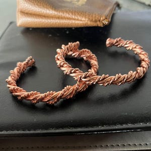 May include: Two copper cuff bracelets featuring a twisted rope design. Each bracelet is constructed from multiple strands of metallic copper wire coiled together to create a textured, continuous spiral pattern with open ends for adjustment.