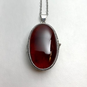 May include: An oval locket pendant featuring a smooth, deep red, translucent stone set in a silver-toned metal frame with a beaded edge. The pendant hangs from a matching silver-toned chain, showcasing a polished finish and a classic design.