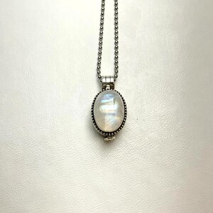 May include: A silver-toned locket pendant featuring an oval-shaped iridescent moonstone cabochon set in a beaded metal frame. The pendant hangs from a matching silver-toned ball chain necklace, showcasing a smooth, reflective surface.