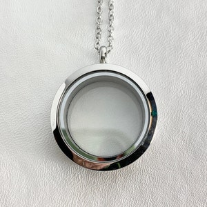May include: A silver-colored, round locket necklace with a clear glass center. The locket hangs from a delicate chain. The locket is empty, ready to be filled with small keepsakes or photos. The background is a textured white surface.