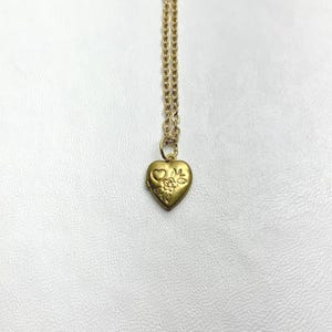 May include: A gold-toned metal necklace featuring a small heart-shaped locket pendant. The locket has a matte finish and is decorated with an embossed design of a single small heart and a flower with leaves on its front surface, attached to a chain.