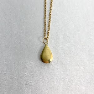 May include: A gold-colored necklace with a teardrop-shaped pendant. The pendant hangs from a delicate chain. The necklace is displayed against a white background.