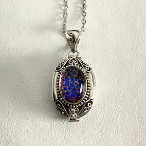 May include: Silver-toned oval pendant necklace with a blue and purple iridescent stone. The pendant has an ornate, filigree design around the stone and a decorative bail. The necklace hangs on a delicate silver chain.