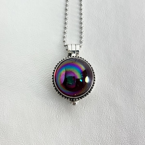 May include: A silver-toned necklace featuring a round pendant with a dichroic glass stone. The stone displays a vibrant array of colors, including blues, greens, and purples. The pendant is encircled by a beaded border and hangs from a delicate ball chain.